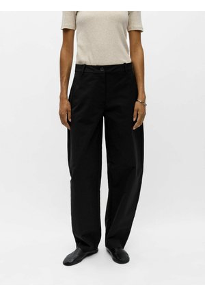 Personne portant un pantalon large noir, des chaussures noires et un haut beige clair à manches courtes, debout devant un fond clair uni.