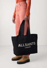 Borsa a tracolla nera in tela con la scritta "ALLSAINTS LONDON" in bianco, dotata di manici robusti e forma piatta. Indossata con jeans rosa e stivali marroni.