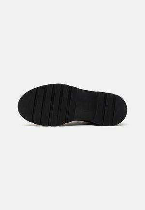 Suela de zapato negra con ranuras profundas horizontales y crestas texturizadas, vista desde abajo sobre un fondo claro y liso.
