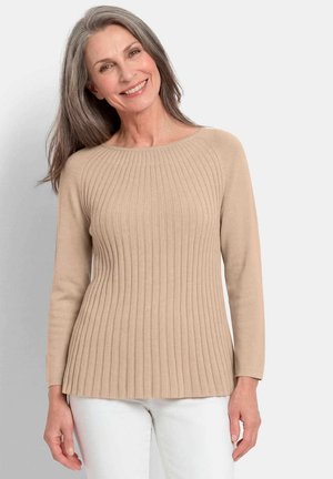 Femme souriante aux longs cheveux gris portant un pull côtelé beige et un pantalon blanc, debout devant un fond clair uni.