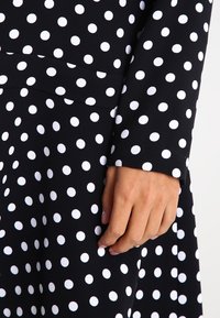 Main reposant sur un tissu noir à pois blancs régulièrement espacés, montrant une partie d'une manche longue et d'une jupe ou robe.