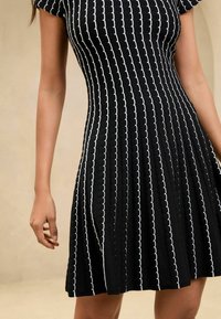 Czarna dzianinowa sukienka z białymi falistymi paskami, krótkimi rękawami, rozkloszowanym dołem i fakturowaną tkaniną. Zaprojektowana, aby podkreślić sylwetkę.