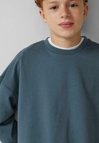 Overmaat, sweatshirt met ronde hals in gedempt blauw katoen. Ribbelboord bij de hals en mouwen. Gelaagd met een witte t-shirt eronder. Glad textuur.