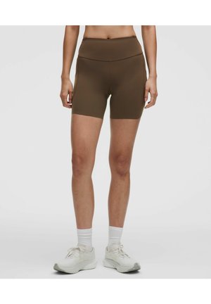 Person iført brune atletikshorts med høj talje, hvide sokker og hvide sneakers, stående på en ensartet lysegrå baggrund.