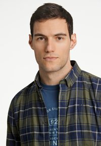 Camisa de franela a cuadros verde y azul marino con collar de botón, superpuesta sobre una camiseta gráfica azul, con un corte casual y tejido texturizado.