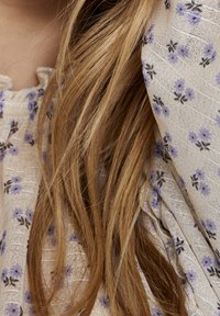 Tissu léger à motif floral avec de petites fleurs violettes et noires, présentant une surface texturée et un drapé doux. Cheveux blonds longs partiellement visibles.