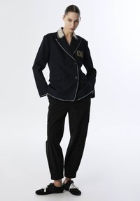 Marineblauwe dubbelgebreide blazer met witte bies en "THE LAB" patch, uitgerust met een gestreepte beige kraag. Gepaarde met zwarte cargobroeken.