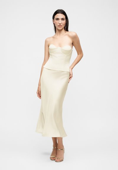 Γυναίκα με μακριά σκουρόχρωμα μαλλιά που φοράει μπεζ κορσέ χωρίς τιράντες και μακριά φούστα στο ίδιο χρώμα, ποζάροντας με το ένα χέρι στη μέση, ουδέτερο φόντο.