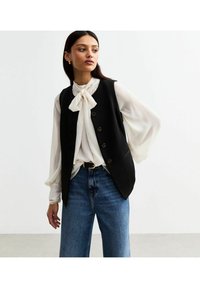 Svart skräddarsydd väst med fem knappar, i kombination med en vit blus med knytkrage. Blå vida jeans fulländar outfiten.