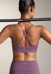 Vijolični športni modrček s prečnim vzorcem na hrbtni strani in prilagodljivimi naramnicami. Material je gladek z diskretnim logotipom na pasu.