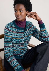 Teal krāsā, ar Fair Isle rakstu un daudzkrāsainiem ģeometriskiem dizainiem, kreklistā ar koka pogām un ribotu apkakli, uzvilkt virs purpura turtlenecka.