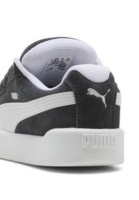 Sneaker in suede grigio con dettagli bianchi. Presenta un colletto imbottito, suola strutturata e un marchio ben visibile su tallone e lato.