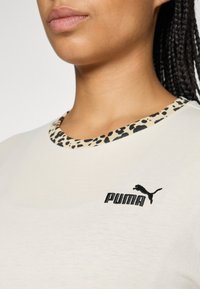 Lähikuva henkilöstä, jolla on yllä valkoinen Puma-paita, jossa on musta logo ja leopardikuosiset pystysuorat kaulukset.