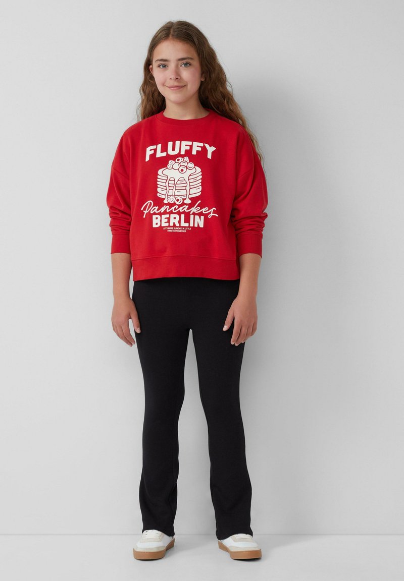 Rode sweatshirt met witte tekst en een pannenkoekafbeelding, gecombineerd met zwarte leggings en witte sneakers met een lichte zool.