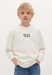 Felpa color crema con collo rotondo e maniche lunghe, caratterizzata da lettere nere a blocco "SCAL PERS" sulla parte anteriore. Indossata con jeans blu.