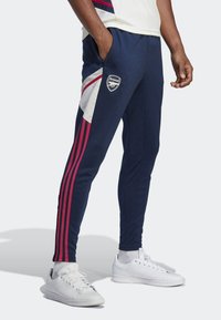 Pantalones deportivos navy con franjas laterales rojas y blancas, que presentan un logo de Arsenal en el muslo y bolsillos con cremallera. Tejido de mezcla de algodón.
