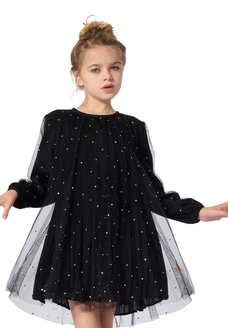 Jeune fille aux cheveux blonds en chignon, portant une robe noire à manches longues avec des points scintillants et un voile transparent, debout, les bras légèrement étendus.