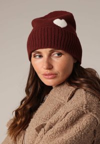 Femme aux longs cheveux bruns portant un bonnet tricoté bordeaux avec un cœur blanc et un manteau beige texturé, regardant sur le côté.