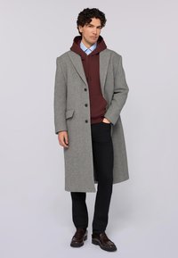 Šedý vlněný kabát s vykrojeným límcem, na dva knoflíky a bočními kapsami, oblečený přes burgundskou mikinu. Černé kalhoty a hnědé boty.