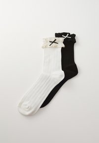 Des chaussettes montantes, une noire et une blanche, avec une texture côtelée. Chacune présente un bord froncé et un accent en croix contrastant dans la couleur opposée.