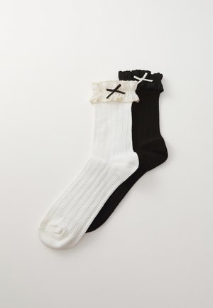 Des chaussettes montantes, une noire et une blanche, avec une texture côtelée. Chacune présente un bord froncé et un accent en croix contrastant dans la couleur opposée.