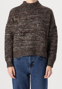 Brauner und grauer melierter Strickpullover mit hohem Kragen und lockerem Sitz, mit überschnittenen Schultern und gerippten Bündchen, kombiniert mit blauen Jeans.