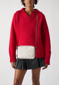 Red knitted sweater with a collar, beige checkered crossbody bag, and a black pleated skirt. Notable details include ribbed cuffs and neutral tones.