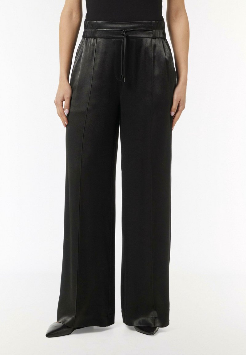 Pantalon en cuir noir à jambes larges avec une taille élastique haute et un cordon, porté avec des chaussures noires pointues et un haut noir.