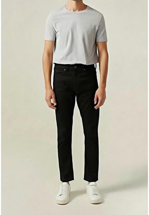 Mann trägt hellgraues, kurz geschnittenes T-Shirt, schwarze, eng anliegende Hose und weiße Sneaker, steht vor einfachem Hintergrund.