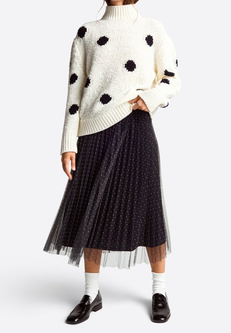 Femme portant un pull à col roulé blanc à pois noirs, une jupe midi plissée noire à petits pois, des chaussettes blanches et des mocassins noirs.