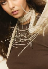 Choker en perles argentées avec des fils délicats, superposé sur un haut ajusté marron. Présente un travail de perles intricate et des accents en tissu transparent.