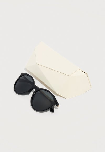 Μαύρα στρογγυλεμένα γυαλιά ηλίου με σκουρόχρωνους φακούς δίπλα σε κλειστή, γωνιακή μπεζ θήκη γυαλιών ηλίου σε απλό ανοιχτόφωτο φόντο.