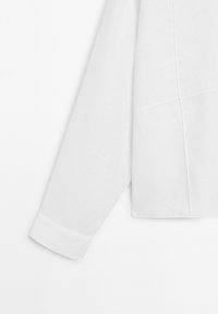 Chemise blanche à manches longues avec une coupe droite, présentant des manches à poignets et un tissu texturé. Les coutures sont visibles pour un détail supplémentaire.