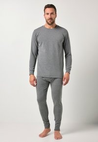 Mann, der barfuß steht und ein enges graues Langarmoberteil sowie passende Leggings trägt, vor einem schlichten hellen Hintergrund.