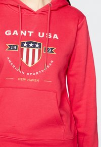 Sweat à capuche rouge avec une poche avant, arborant un logo en forme de bouclier avec des rayures et des étoiles, ainsi que les textes "GANT USA" et "AMERICAN SPORTSWEAR."