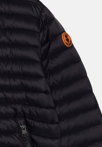 Giacca a vento nera con texture trapuntata, dotata di una patch arancione con logo sulla spalla e di una zip nera all'orlo.