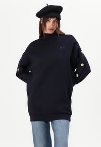Navy oversized sweatshirt made of soft fabric with high collar, embroidered logo, and gold button accents on sleeves. Paired with denim.