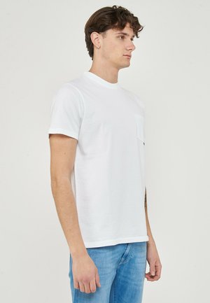 Giovane uomo con capelli castani corti che indossa una t-shirt bianca a maniche corte e jeans azzurri, in piedi di profilo su uno sfondo neutro.