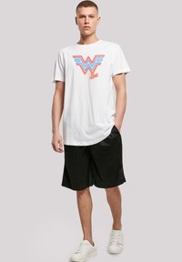 Hvit bomulls t-skjorte med blå og rød grafikk "WW" logo; kombinert med svarte mesh-shorts og hvite joggesko. Enkelt design, avslappet passform.