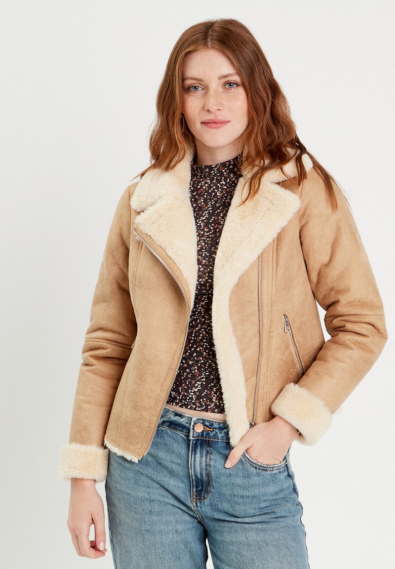 Chaqueta de ante sintético marrón con cuello y puños de borreguito, cierre delantero con cremallera y bolsillos laterales. Llevada sobre una blusa de estampado floral y unos vaqueros.