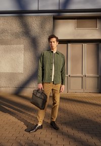 Veste verte à boutons, chemise beige, pantalon couleur tan, chaussures noires et une mallette en cuir noire ; debout contre un mur texturé.