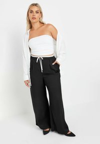 Svarta vida byxor med vit dragsko i midjan, kombinerat med en figursydd vit strapless topp och en öppen vit skjorta. Spetsiga svarta skor.