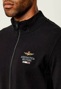 Felpa nera con zip, caratterizzata da un colletto alto, logo ricamato con accenti dorati e strisce di colore contrastante sul fondo.