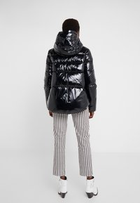 Osoba ubrana w błyszczącą czarną kurtkę puchową z kapturem, w paskowane spodnie do kostek oraz białe botki na obcasie, stojąca na jednolitym tle.