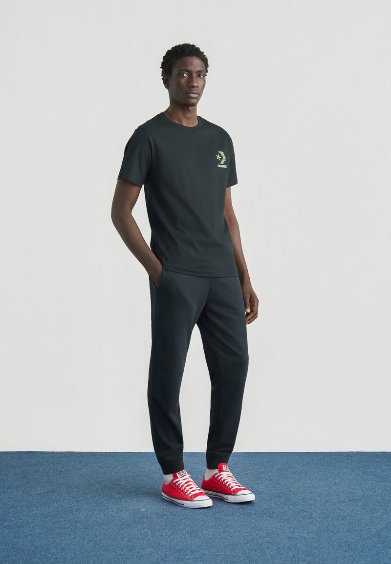 T-shirt nera in cotone con una piccola grafica verde, abbinata a pantaloni da jogging scuri e sneaker Converse rosse. Sfondo semplice.
