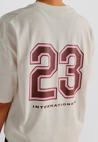 Flødefarvet t-shirt med et stort brunt og pink tal "23" og ordet "INTERNATIONALS" nedenunder i sort. Kortærmet, klassisk pasform.