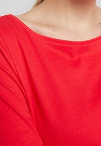 Nærbillede af en person iført en lys rød langærmet skjorte med en bred, rund halsudskæring.