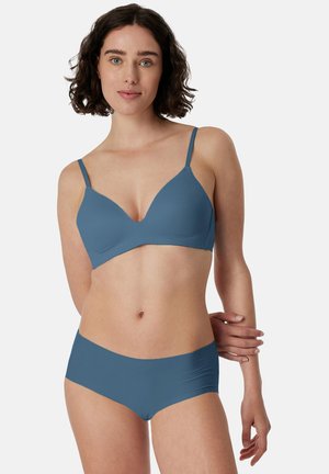 Jeune femme aux cheveux courts et bouclés portant un soutien-gorge et un shorty bleus assortis, debout devant un fond clair uni.