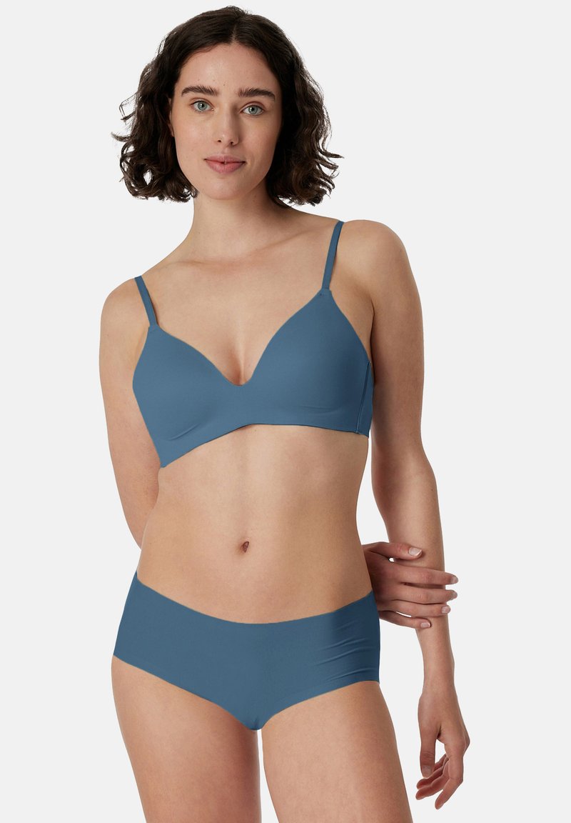 Giovane donna con capelli corti e ricci che indossa un reggiseno e slip blu senza cuciture abbinati, in piedi contro uno sfondo chiaro e uniforme.