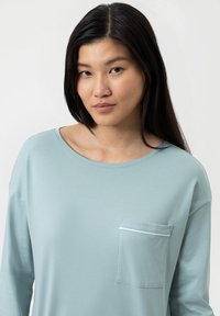 Un haut bleu clair à manches longues en tissu doux, avec un col arrondi et une petite poche poitrine ornée d'une discrète bordure blanche.